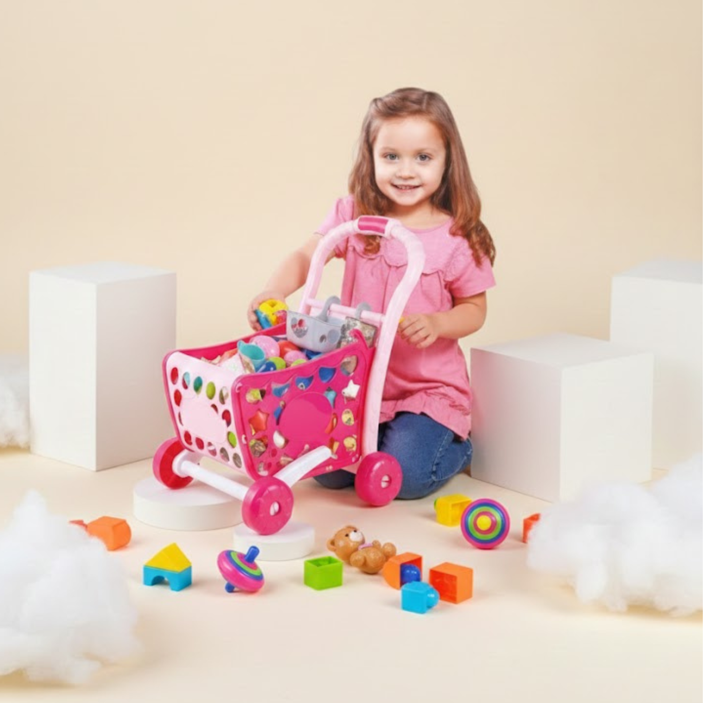 Little girl playing with a pink toy shopping cart filled with colorful pretend play accessories.
